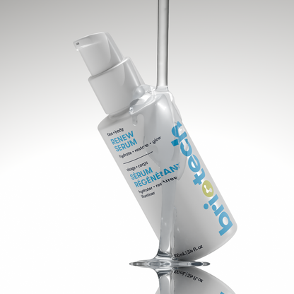 A white bottle of Renew Serum by britech, with a pump dispenser, stands upright on a reflective surface, partially wrapped in splashing clear liquid. The label displays blue and gray text.